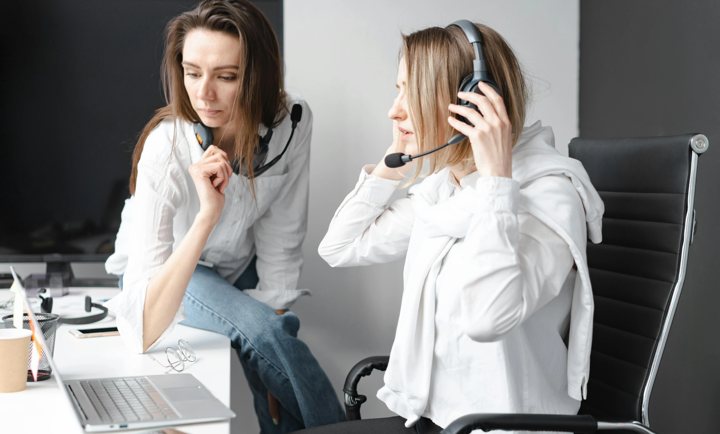 Two customer service agents wearing headsets discuss information at a desk with a laptop.
