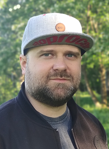 A portrait of me wearing a gray cap and a black bomber jacket, standing among green trees.