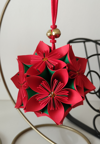 Red and green paper flower ball ornament with a gold bell on a ribbon, hung on a gold stand.